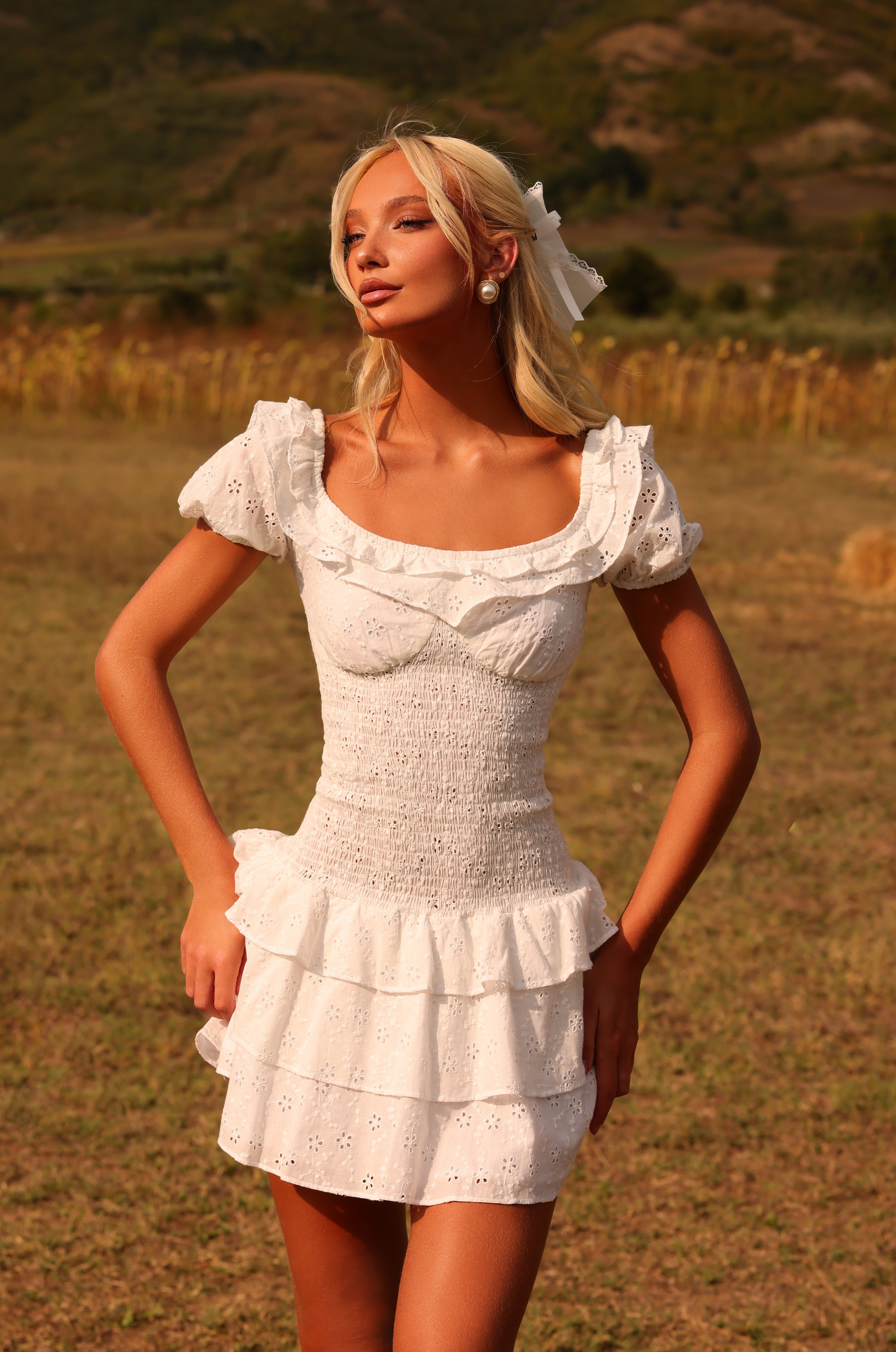 A woman in the Ivory White Cotton Mini Graduation Dress: Macie stands outdoors in a sunlit, rural landscape.